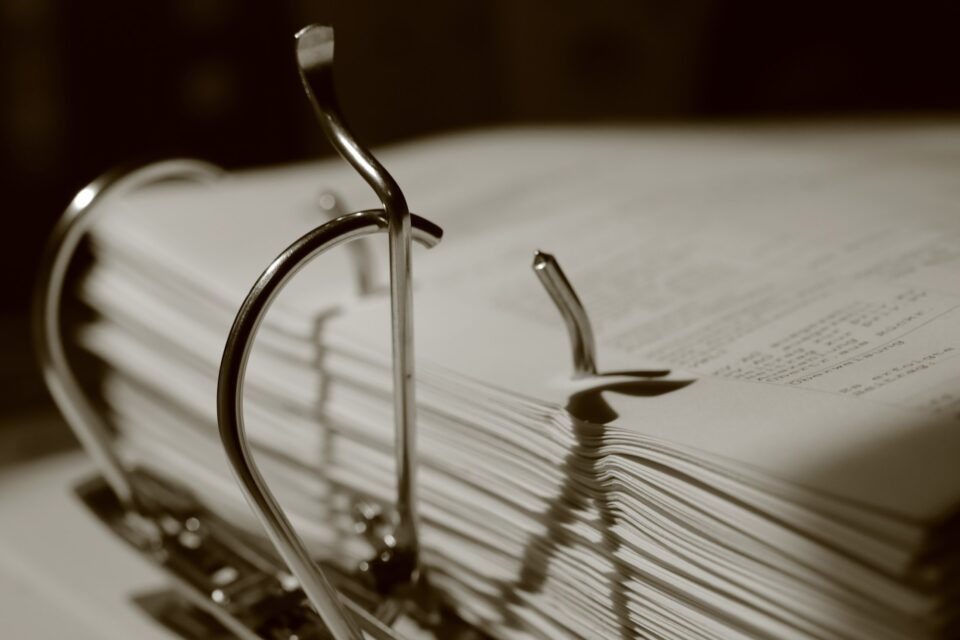 Muted editorial close-up of a thick stack of paper records secured by metal binder rings, representing structured support requests